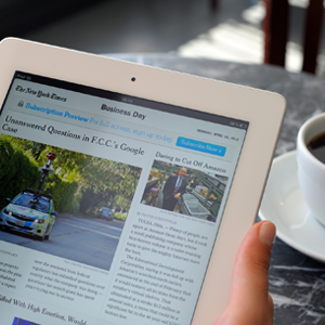 2 mains tiennent une tablette qui affiche une revue; une tasse de café est visible en second plan.